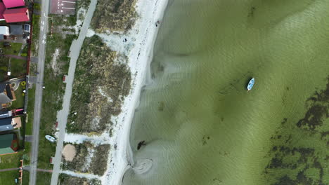 Die-Luftaufnahme-Zeigt-Ein-Einsames-Boot,-Das-In-Seichtem,-Grünem-Wasser-In-Der-Nähe-Eines-Weißen-Sandstrands-Schwimmt,-Der-Von-Natürlichem-Schutt-Gesäumt-Ist,-Im-Kontrast-Zu-Leuchtend-Rosa-Gebäuden-Und-Einer-Belebten-Straße