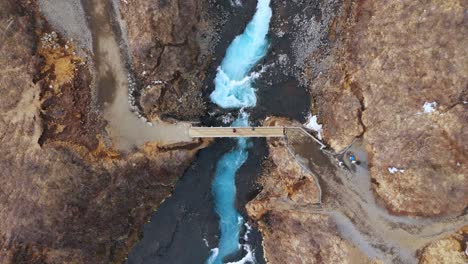 Bruarfoss-waterfall-in-iceland,-with-a-bridge-over-vibrant-blue-glacial-waters,-aerial-view