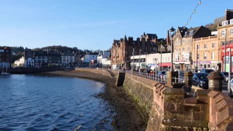 Scenic-Scottish-view-of-Oban-town-with-people-walking-and-traffic-driving-along-waterfront-on-a-blue-sky-sunny-day-in-Western-Scotland-UK