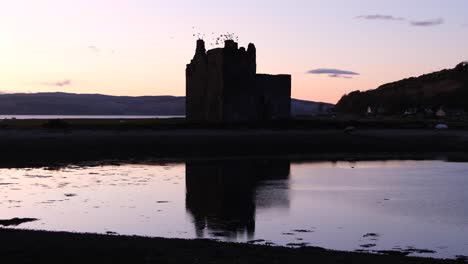 Lochranza-Castle-silhouetted-during-twilight-sunset-with-reflection-onto-ocean-water-on-Isle-of-Arran,-with-flock-of-birds-flying-overhead-in-Western-Scotland-UK