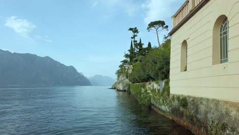 Explore-El-Encanto-Del-Lago-De-Garda-Desde-Malcesine,-Capturando-Su-Belleza-Arquitectónica-Entre-Frondosos-árboles,-Aguas-Azules-Y-Cielos-Pintorescos.