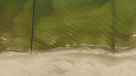 Top-down-aerial-view-of-a-serene-beach-with-clear-green-sea-waters-gently-lapping-against-a-sandy-shoreline,-marked-by-subtle-wave-patterns-and-a-single-trail-across-the-sand