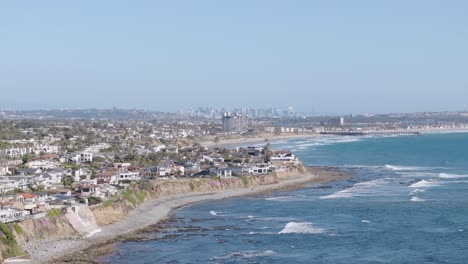 Aerial-track-back-of-San-Diego,-California-shore-and-cliffside-neighborhood-with-the-skyline-in-the-background-on-a-sunny-day