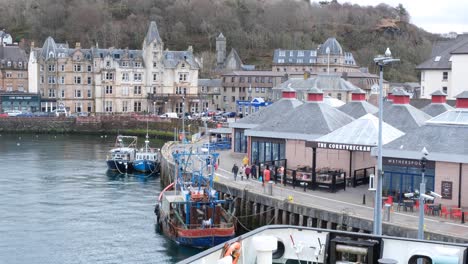 People,-tourist,-visitors,-and-locals,-walking-alongside-the-harbour-waterfront-with-cafes-and-pubs,-and-moored-boats-and-ships-in-Oban-town,-Western-Scotland-UK