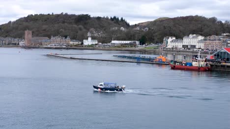 Local-fishing-tour-boat-heading-out-of-Oban-bay-and-harbour-with-town,-houses,-and-hotels-in-the-distance-of-Western-Scotland-UK