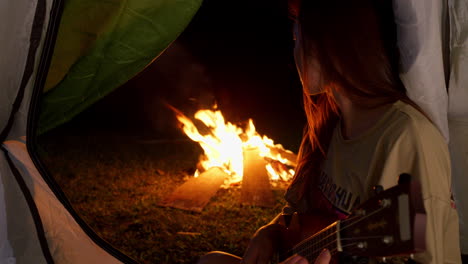 close-up-of-young-asiatic-traveller-playing-alone-ukulele-guitar-in-front-of-bonfire-at-night-inside-her-camping-tent