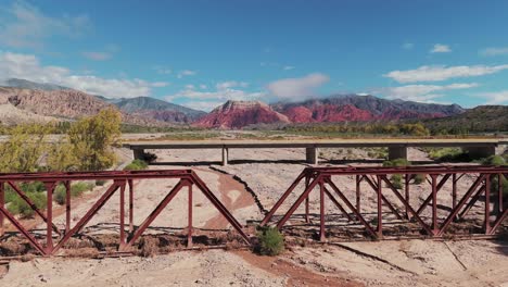 Drone-flying-over-a-railway-bridge-and-a-car-bridge-on-Route-9-in-Jujuy,-Argentina,-with-a-breathtaking-backdrop-of-colorful-mountains