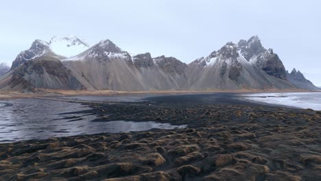 Schwarzer-Sandstrand-Bei-Ebbe-Mit-Schneebedeckter-Bergkette-Dahinter