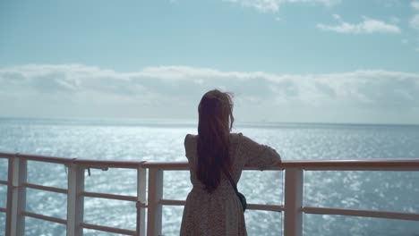 Frau-In-Einem-Kleid-Auf-Dem-Deck-Eines-Kreuzfahrtschiffs-Am-Mittag,-Spürt-Den-Wind-Und-Blickt-Auf-Das-Meer,-Rückaufnahme-Langsam-Lang