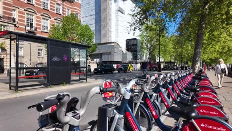 Row-Of-Empty-Parked-Santander-hire-bikes-stationed-at-Westminster-With-Cyclists-Going-Past-In-Background