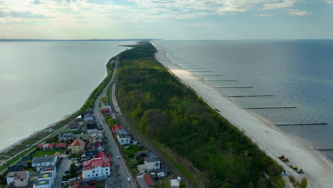 Aerial-view-of-Kuźnica,-Pomorskie,-thin-strip-of-land-dividing-the-Baltic-Sea-from-the-bay,-lined-with-a-lush-green-forest,-residential-areas,-and-a-sandy-beach-stretching-into-the-distance