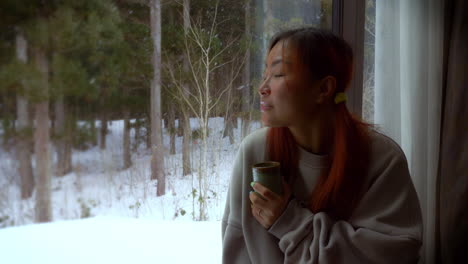 Beautiful-Girl-leaning-by-a-window,-looking-out-at-a-snowscape,-snowing-and-trees-in-the-background