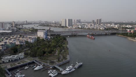 Ho-Chi-Minh,-Saigon,-Vietnam,-with-the-river,-a-bridge,-and-a-cargo-vessel-passing-under-it,-aerial-drone-video-during-sunset