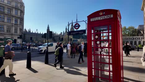 Passersby-leisurely-walk-past-an-iconic-red-telephone-box-near-the-entrance-of-Westminster-Station-on-Parliament-Street,-London,-embodying-the-concept-of-urban-charm-and-cultural-heritage