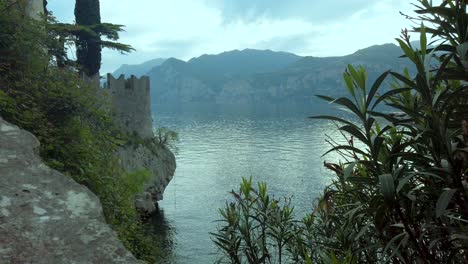 üppige-Vegetation-Am-Seeufer,-Umrahmt-Von-Ruhigem-Wasser,-Hohen-Bergen-Und-Einem-Weiten-Himmel-Am-Gardasee