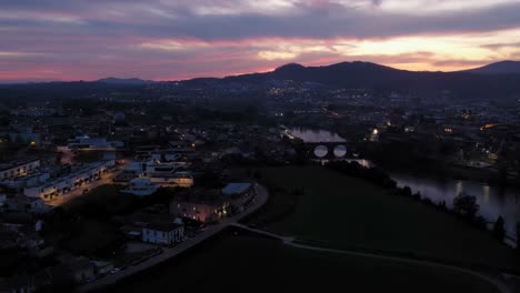 Luftaufnahme-Von-Barcelos-In-Der-Abenddämmerung-Mit-Beleuchteten-Straßen-Und-Einer-Alten-Brücke-über-Den-Fluss,-Vor-Einem-Lebhaften-Sonnenuntergangshimmel