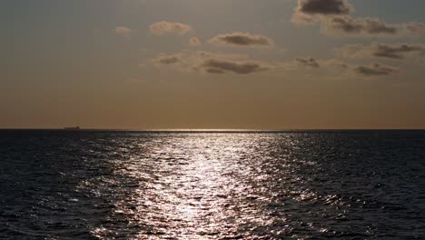 Neigung-Nach-Unten-Von-Der-Sonne-Hoch-Am-Himmel-Mit-Tanker-Am-Horizont,-Während-Sonnenlicht-Auf-Dunklem-Wasser-Glitzert