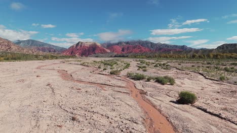 Drone-flying-over-a-dry-river-in-summer-with-beautiful-colorful-mountains-in-the-background