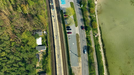 Aerial-view-capturing-the-juxtaposition-of-a-railway-and-a-road-running-parallel-along-a-lush-green-shoreline,-with-vehicles-and-a-train-visible,-showcasing-infrastructure-nestled-in-a-natural-coastal