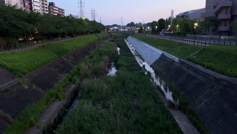 Dusk-descends-on-a-serene-urban-drainage-canal-surrounded-by-lush-greenery,-shot-from-an-aerial-perspective