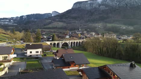 Schöne-Weiße-Backsteinbrücke-über-Den-Fluss-Mit-Einer-Kleinen-Französischen-Stadt-Und-Bergen-Im-Hintergrund-Und-Einem-Grünen-Rasen-Davor