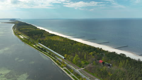 Aerial-view-capturing-a-train-traveling-along-the-narrow-Hel-Peninsula-in-Kuźnica,-Poland,-dividing-the-tranquil-bay-from-the-vast-Baltic-Sea