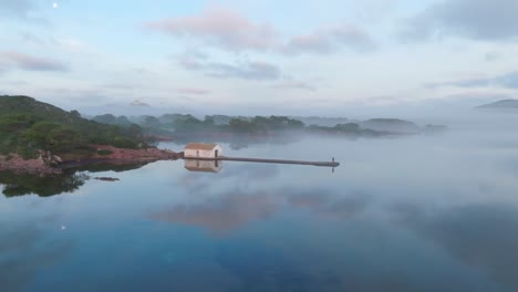 Nubes-Espejadas-Reflexión-Sobre-La-Bahía-Islote-De-Aguas-Tranquilas-Acantilado-Verde-Costa-Drone-Estableciendo-Toma-Aérea