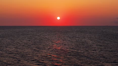 Aerial-pullback-above-calm-ocean-tropical-waters-as-red-orange-yellow-ball-of-sun-sets