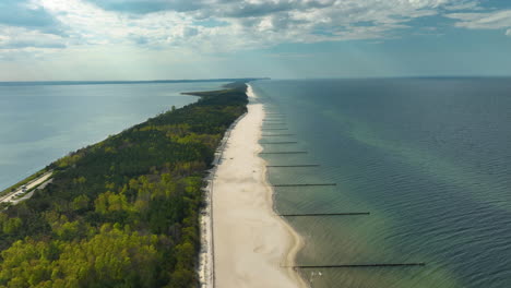 Aerial-view-showcasing-the-slender,-forest-fringed-Kuźnica-peninsula,-with-a-clear-separation-between-the-serene-Baltic-Sea-and-the-tranquil-bay,-highlighted-by-groynes-extending-into-the-sea
