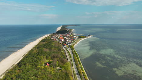 Aerial-view-of-Kuźnica,-showing-a-vibrant-village-nestled-between-the-greenish-blue-waters-of-the-bay-and-the-deep-blue-sea,-bisected-by-a-railway-and-lush-greenery