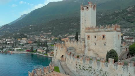 Aerial-view-revealing-Malcesine-Castle-and-its-medieval-village-nestled-amidst-water,-sky,-and-mountains