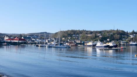 Pan-of-Scottish-Oban-town,-harbour-and-beach-with-moored-ships-and-boats-on-a-sunny-blue-sky-day-in-popular-tourism-destination,-Western-Scotland-UK