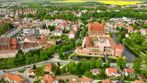 This-aerial-view-captures-the-extensive-layout-of-Lidzbark-Warmiński's-castle,-set-against-a-backdrop-of-the-town's-varied-architectural-styles,-greenery-and-a-serene-river-curving-around-the-castle