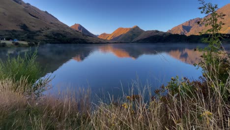 Mountains-and-blue-sky-reflecting-off-calm-water-at-Moke-Lake-with-early-morning-fog