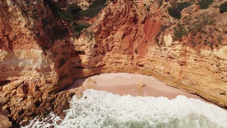 Top-down-view-of-Coastline-waves-crashing-with-pan-up-to-orange-rugged-cliffs-by-aerial-4k-drone-in-the-Algarve-region-of-Portugal