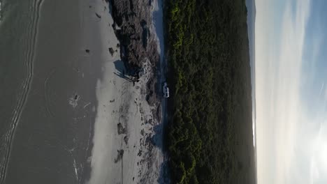 Vehicle-traveling-near-the-seashore-with-white-sand-in-Wylie-Bay-Rock-Beach,-vertical-aerial
