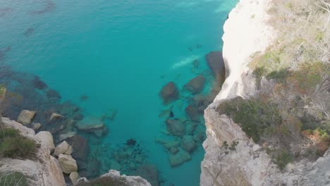 Aerial-Drone-Tops-down-cliff-at-blue-turquoise-water-beach-in-Menorca-Forested-coastal-seaside-area,-natural-environment-travel-destination