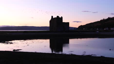 Scenic-landscape-view-of-Lochranza-Castle-on-Isle-of-Arran,-silhouetted-during-twilight-sunset,-with-flock-of-birds-flying-over-historical-landmark,-Western-Scotland-UK