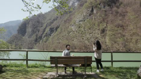 Mujer-Tomando-Fotografías-De-Un-Hombre-Comiendo-En-El-Banco-Del-Parque-Junto-Al-Lago.