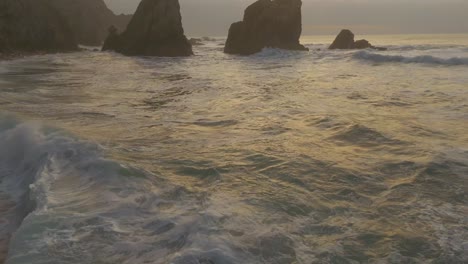 Aerial-view-tilting-over-waves-on-Atlantic-Ocean-at-the-Magoito-Beach,-cloudy-sunset-in-Portugal