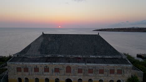 Old-world-charm-yellow-abandoned-building-overlooks-ocean-revealing-setting-sun-on-horizon,-drone