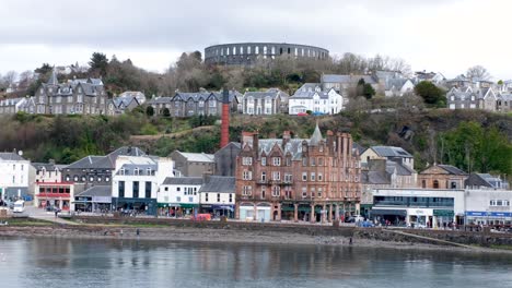 Malerische-Landschaftsansicht-Der-Stadt-Oban-Mit-Menschen-Und-Verkehr-Entlang-Der-Hafenpromenade-Und-McCaig&#39;s-Tower-Auf-Hügeln-Im-Westen-Schottlands,-Vereinigtes-Königreich