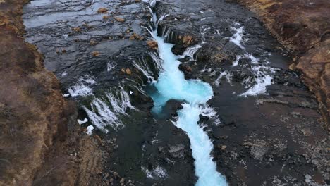 Icy-Bruarfoss-Waterfall-cutting-through-brown-Icelandic-terrain,-aerial-view