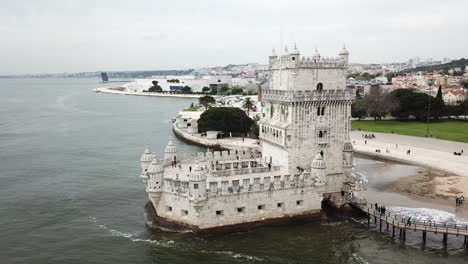 Lisbon,-Belem-tower's-drone-footage-zooming-in-from-the-right-hand-side-of-the-tower-where-the-pedestrian-bridge-and-the-river-can-be-seen-as-well-as-the-buildings-settlement-behind-the-tower