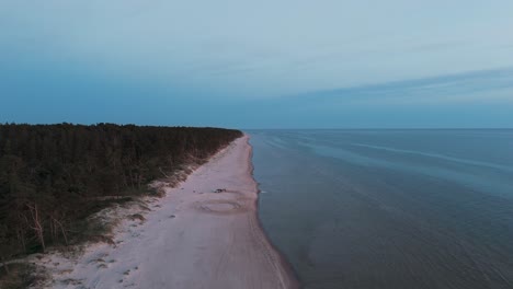 Car-Stuck-in-Sand-at-the-Dune-Sea-Coast-of-Baltic-Sea-Bernati,-Latvia-Sun-Going-Beyond-the-Horizon-Illuminating-Water-and-Clouds-in-the-Sky,-Calm-Sea-Waves,-Romantic-Mood,-Wide-Angle-Drone-Shot