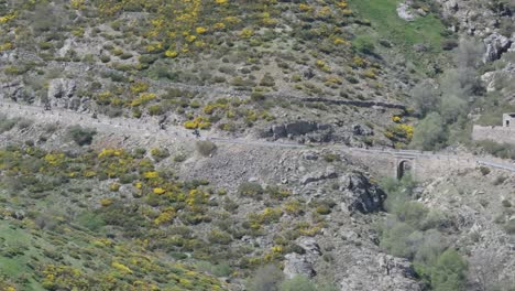 Drone-filming-of-a-mountain-road-where-we-see-bikers-driving-downhill-one-after-the-other,-passing-over-a-bridge-and-next-to-a-large-stone-house-in-ruins-in-Sierra-de-Gredos,-Spain