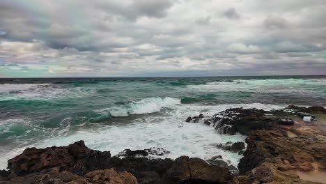 Waves-roll-onto-a-rocky-shoreline,-displaying-shades-of-turquoise-set-against-a-cloudy-sky,-highlighting-the-pristine-water-quality