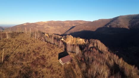 Aerial-parallax-drone-shot-of-a-wooden-cabin-surrounded-by-sparse-trees-on-a-sunny-autumn-day,-clear-sky,-in-the-apuseni-area,-Romania
