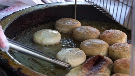 El-Pastel-De-Carne-Chino-Recién-Preparado-Chisporrotea-En-Una-Sartén-De-Aceite-Caliente-Mientras-Un-Vendedor-Voltea-Hábilmente-La-Masa-Con-Pinzas-De-Cocina-Y-La-Fríe-Hasta-Que-Esté-Dorada,-Primer-Plano-De-La-Popular-Comida-Asiática