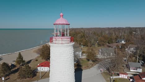 A-white-lighthouse-with-a-red-top-on-a-sunny-day,-overlooking-a-beach-and-a-quiet-neighborhood,-aerial-view